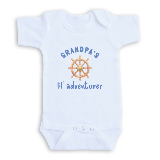 A white baby onesie with a graphic of a ship's steering wheel and the text "GRANDPA'S lil' adventurer" printed on it.