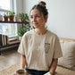 A woman wearing a beige t-shirt with text and a coffee cup is sitting on a couch in a room with potted plants and bookshelves.