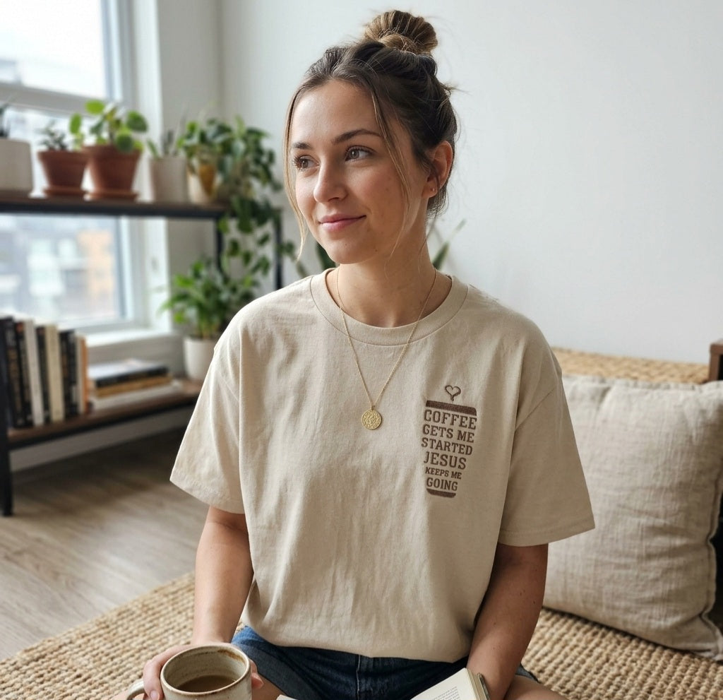 A woman wearing a beige t-shirt with text and a coffee cup is sitting on a couch in a room with potted plants and bookshelves.