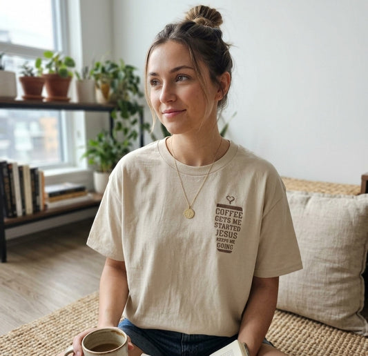 A woman wearing a beige t-shirt with text and a coffee cup is sitting on a couch in a room with potted plants and bookshelves.