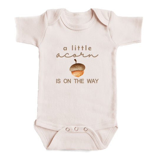 A baby onesie with the text "a little acorn is on the way" printed on it.