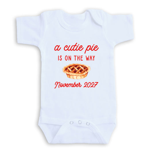 A white baby onesie with a red text and graphic design that reads "a cute pie is on the way November 2027".