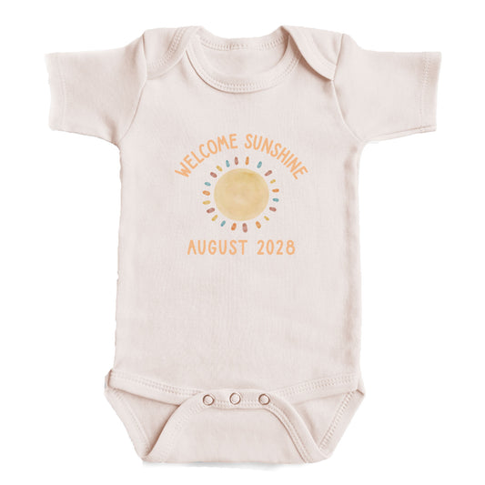 A baby onesie with a sun design and text that reads "Welcome sunshine August 2028".