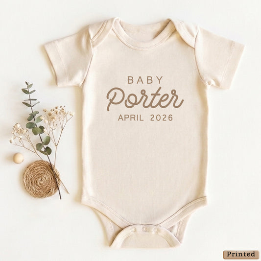 A baby onesie with the text "BABY Porter APRIL 2026" printed on it, along with some dried flowers and twigs.