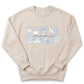 A beige sweatshirt with a graphic design featuring various objects such as a teapot, cat, butterfly, and other decorative elements arranged in a row.