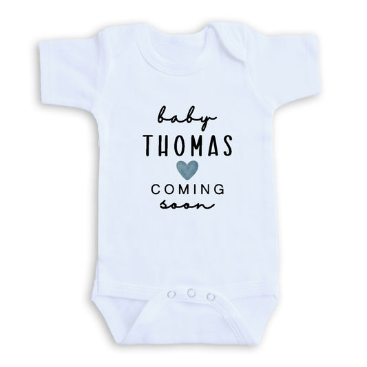A white baby onesie with the text "baby Thomas coming soon" printed on it.