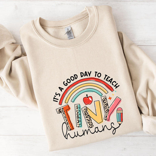 A beige sweatshirt with a colorful graphic design on it, which says "It's a good day to teach tiny humans".