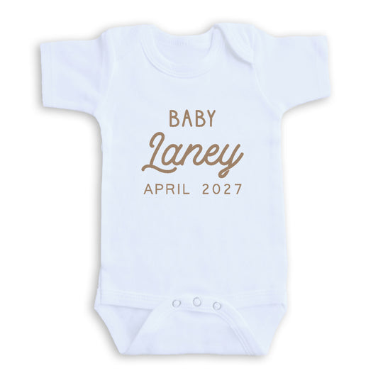 A white baby onesie with the text "BABY Laney April 2027" printed on it.