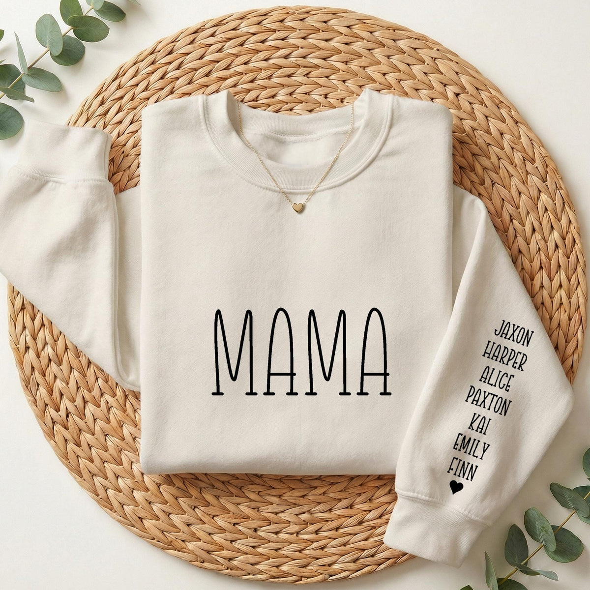 A white long-sleeved shirt with the word "MAMA" printed on it, along with names below the text, is displayed on a woven placemat.
