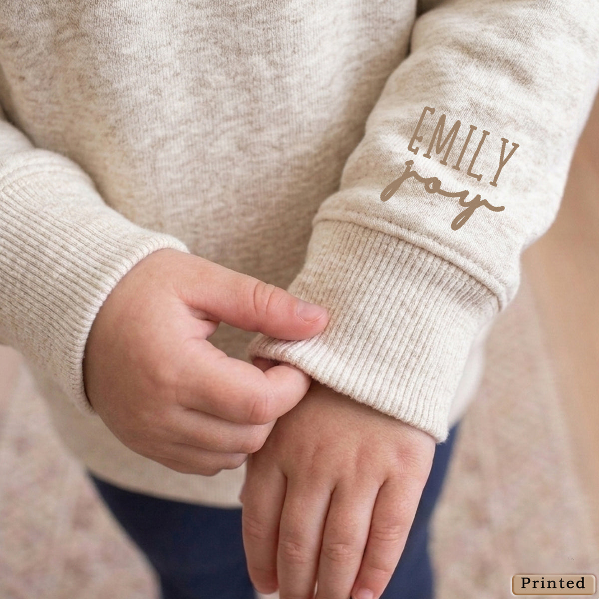 A close-up of a person's hand wearing a beige sweater with the name "Emilly" printed on the sleeve.