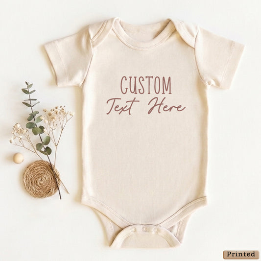 A beige baby onesie with the text "CUSTOM Text Here" printed on it, along with some dried flowers and twigs.