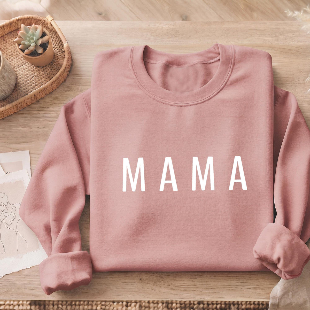A pink sweatshirt with the word "MAMA" printed on it, placed on a wooden surface alongside a small potted plant and some papers.