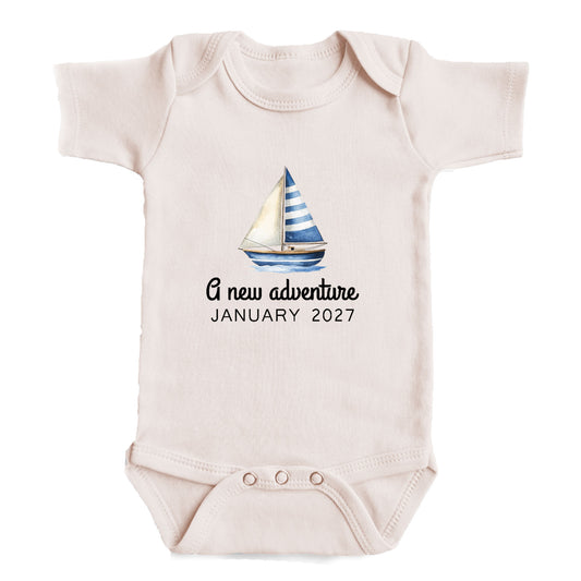 A baby onesie with a sailboat graphic and the text "A new adventure JANUARY 2027" printed on it.