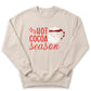 A beige sweatshirt with a red and white design featuring a Santa Claus mug and the text "HOT COCOA SEASON" printed on it.